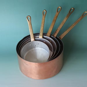 May include: A set of five nested saucepans with polished copper exteriors and silver-toned metal interiors. Each pan features a long, slender brass handle with a teardrop-shaped hole at the end for hanging, attached by three visible rivets.