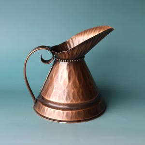 May include: A copper-coloured metal pitcher with a hammered texture, featuring a wide, flared spout and a curved handle. The rim of the pitcher is decorated with a beaded detail, and the body has a conical shape that tapers towards the top.