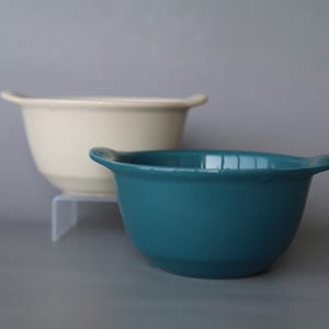 May include: Two ceramic bowls with side handles, one in a solid cream colour and one in a solid teal colour. The bowls feature a smooth, rounded design with a wide opening and a tapered base, presented in a stacked arrangement for clear visibility.