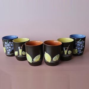 May include: A set of six dark brown ceramic tumblers with hand-painted designs. Two cups feature blue grapes, two feature yellow lemons, and two feature glasses of lemonade. The interiors are colored in light blue, yellow, and orange tones.