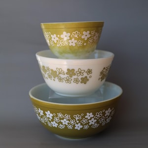 May include: A stack of three glass mixing bowls. The top bowl is olive green with white floral patterns. The middle bowl is white with olive green floral patterns. The bottom bowl is olive green with white floral patterns. All bowls are round.