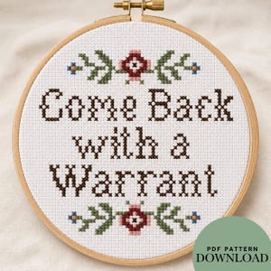 May include: A circular wooden embroidery hoop containing white Aida fabric with the cross-stitched text Come Back with a Warrant in dark brown. The design features symmetrical floral motifs in green, pink, and blue at the top and bottom.