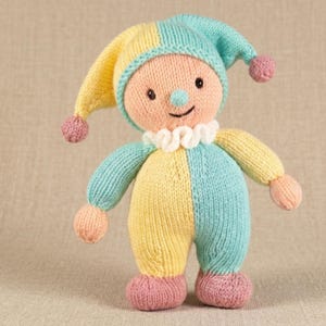 May include: A knitted doll featuring a jester-style hat with two pom-poms, a peach-coloured face, and a ruffled white collar. The body is constructed with alternating panels of pastel yellow and light blue yarn, finished with pink feet.