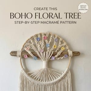 May include: A macramé wall hanging featuring a circular wooden hoop and a horizontal piece of driftwood. The design depicts a tree with off-white cord knots and small, colorful crocheted flowers in pink, blue, yellow, and purple hues.