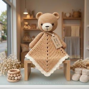 May include: A tan crochet teddy bear lovey blanket with a cream-colored muzzle, ears, and scalloped border. A beige ribbon holds a rectangular tag that reads Teddy Lovey Blanket - $65. The bear has dark brown eyes and a stitched black nose.