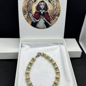 May include: A bracelet featuring alternating light green round beads and light tan cylindrical wooden beads, secured with a silver-toned metal lobster clasp, presented inside a white box with a circular illustration of a hooded figure.