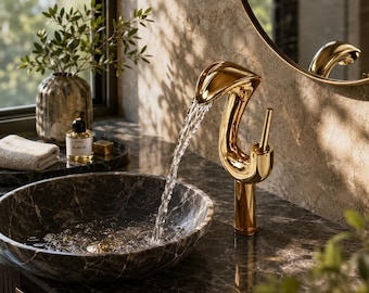 May include: A polished gold-toned faucet with a curved, sculptural design dispensing a steady stream of water into a dark, circular marble vessel sink. The faucet features a slender vertical handle and is set against a matching marble countertop.