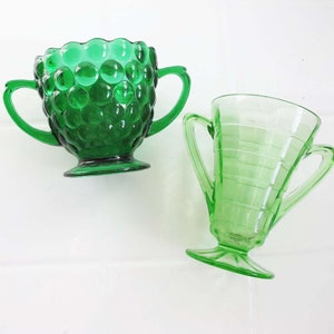 May include: Two green glass sugar bowls with handles. One bowl has a textured, bubbled surface. The other bowl has a smooth, ribbed surface.