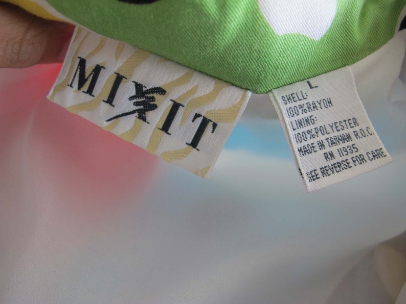 May include: A white, blue, and pink rayon jacket with a "MIXIT" label and a size L tag. The tag lists the fabric composition as 100% rayon for the shell and 100% polyester for the lining. It also states that the jacket was made in Taiwan and includes the RN number 11935. The tag instructs the user to see the reverse side for care instructions.