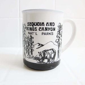 May include: A white ceramic mug with a black handle and base. The front features a black line drawing of a bear, trees, and mountains with the text "SEQUOIA AND KINGS CANYON NAT'L PARKS" printed in a bold, stylized black font above the illustration.