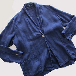 May include: A navy blue linen blazer with a single-button closure and two front pockets. The long-sleeved jacket has a shawl collar and buttoned cuffs. The garment is made of a textured fabric.