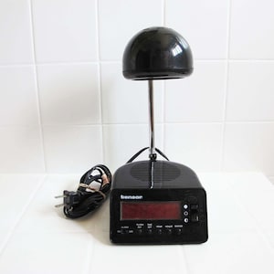 May include: A black Tensor alarm clock with a digital display and a distinctive design. It has a dome-shaped top and a silver stem. The clock includes function buttons and a power cord.