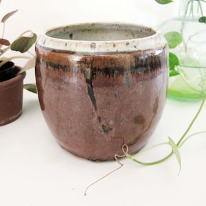 May include: A handcrafted, brown ceramic planter with a wide, rounded shape and a cream-colored rim. The planter has a glossy finish and a dark brown, almost black, vertical streak. A small potted plant and a glass vase with greenery are in the background.