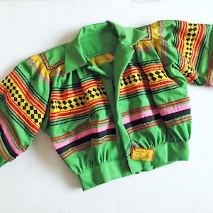 May include: Green jacket with colourful striped and chequered patterns in red, yellow, black, and pink. The jacket has a collared neckline and long, gathered sleeves. A small tag is sewn on the front.