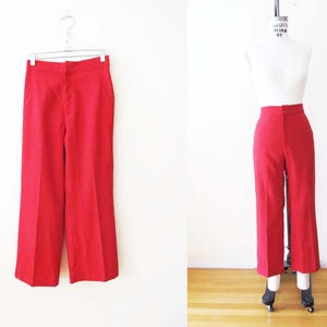 May include: Bright red, high-waisted pants. The pants are displayed on a hanger and a mannequin. The pants have a straight leg design and a button and zipper closure. The fabric appears to be a smooth, woven material.