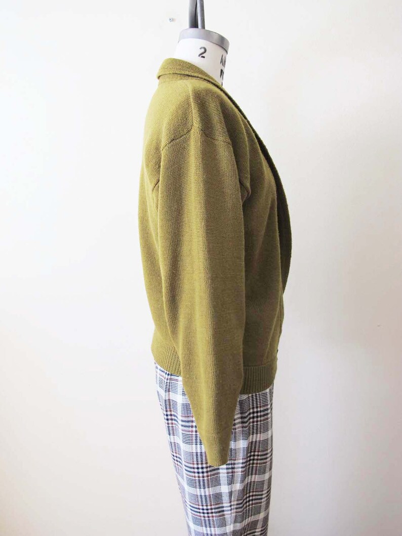 Puede incluir: Un c&aacute;rdigan de manga larga de color verde oliva con cuello. El c&aacute;rdigan est&aacute; en un maniqu&iacute; y las mangas est&aacute;n remangadas.