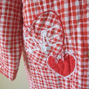 May include: Red and white gingham quilted robe with a large red heart patch on the front. The heart has white stitching around the edges and a white bow in the center.