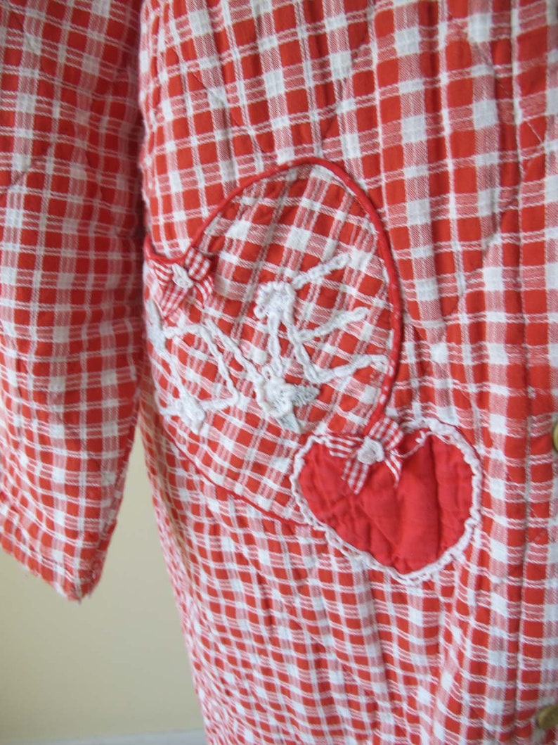 May include: Red and white gingham quilted robe with a large red heart patch on the front. The heart has white stitching around the edges and a white bow in the center.