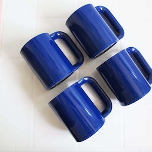 以下が含まれることがあります： 白いタイルの上に配置された、大きなハンドルが付いたコバルトブルーのセラミックマグ4つ。 マグカップは均等に配置され、角度がつけられており、光沢のある仕上げとシンプルなデザインが特徴です。 ピンク色の背景が見えます。