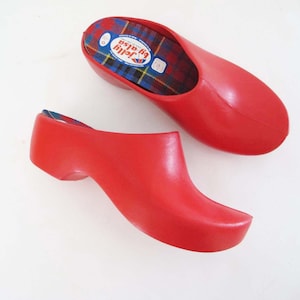 May include: Two bright red clogs with a classic design. The clogs have a rounded toe and a low heel. The interior features a plaid pattern. The clogs are made of a smooth, solid material. The brand name "Jolly by alsa" is visible inside one clog.