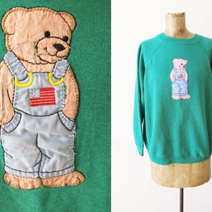 Puede incluir: Sudadera verde con un osito de peluche rosa que lleva un mono y un parche de la bandera estadounidense bordado en la parte delantera.