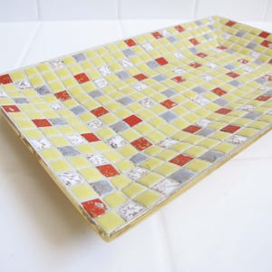 May include: A rectangular tray featuring a mosaic surface composed of small square tiles in shades of pale yellow, vibrant red, and metallic silver. The tiles are arranged in a grid pattern across the flat interior surface of the tray.