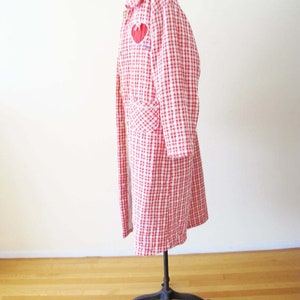 May include: A red and white gingham quilted coat with a red heart embroidered on the front. The coat is on a mannequin and has long sleeves and a collar.