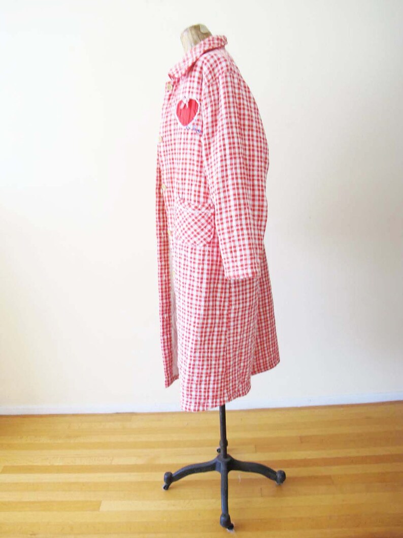May include: A red and white gingham quilted coat with a red heart embroidered on the front. The coat is on a mannequin and has long sleeves and a collar.