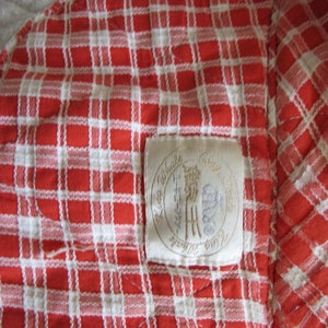 May include: A red and white checkered fabric with a white label sewn into the corner. The label has the text "Little Village" and a logo with a stylized "H" inside a circle.