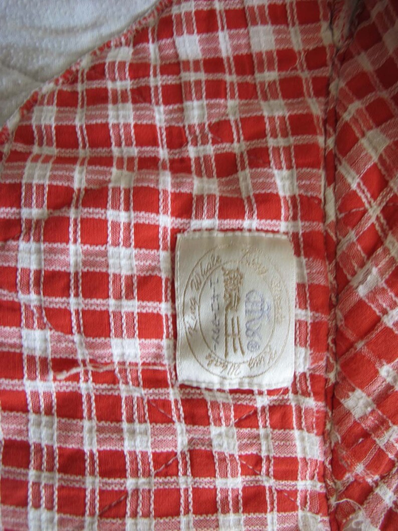 May include: A red and white checkered fabric with a white label sewn into the corner. The label has the text "Little Village" and a logo with a stylized "H" inside a circle.