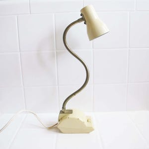 Puede incluir: Una lámpara de escritorio vintage con una pantalla y base de color crema. El brazo flexible de cuello de cisne es de color plateado metálico. La lámpara está enchufada a una toma de corriente blanca. La lámpara está sobre una superficie blanca.