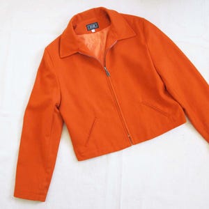 May include: An orange jacket with a collar and a full-length zipper. The jacket has two front pockets and a small label near the collar. The jacket is made of a smooth fabric and is a solid color.