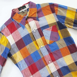 Può includere: Una camicia patchwork a maniche lunghe in una varietà di colori, tra cui rosso, blu, giallo e arancione. La camicia presenta un colletto, una tasca sul petto e chiusure con bottoni. Il design patchwork è composto da quadrati.