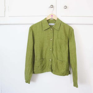May include: A long-sleeved, olive green linen shirt with a collared neckline and button-down front. The shirt has two front pockets and is hanging on a wooden hanger against a white background.