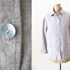 May include: A light gray linen shirt with a button-up front and long sleeves. The shirt has two pockets on the front and a mother-of-pearl button.