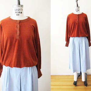 Puede incluir: Una camisa de manga larga de color óxido con cuello abotonado se combina con pantalones plisados ​​de color azul claro. La camisa tiene cuello redondo y puños ajustados. El conjunto se exhibe en un maniquí.