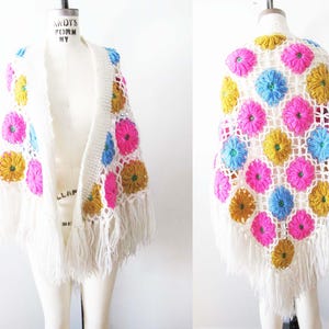 May include: A white crochet shawl with a floral pattern in pink, blue, and yellow. The shawl has a fringe along the bottom edge and is displayed on a mannequin. The open weave design is visible.