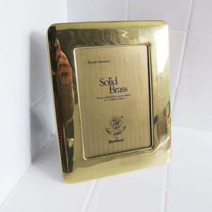 May include: A rectangular, solid brass photo frame with rounded corners. The frame has a polished, reflective surface and features the text "Solid Brass" and "Tarnish Resistant". The frame measures 5" x 7" (12.7cm x 17.78cm).