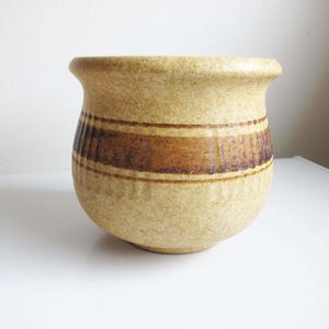 May include: A beige ceramic pot with a rounded body and a slightly flared rim. The pot features a textured surface and a horizontal band of brown stripes around the middle. The pot is likely used for decorative purposes or as a planter.