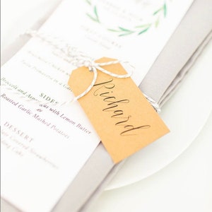 May include: A brown paper place card with the name "Richard" written in cursive on it. The place card is tied with twine and is sitting on top of a folded gray napkin.