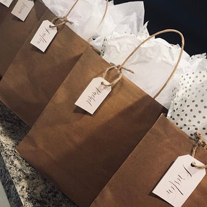 May include: Four brown paper gift bags with white tissue paper and personalized tags. The tags are tied with twine and have names written in calligraphy.
