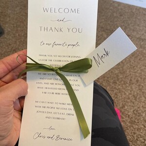 May include: A white wedding program with a green ribbon tied around it. The program says "Welcome and Thank You to our favorite people." and "We are so blessed to have each and every one of you in our lives, and are honored that you have traveled from near and far to be here with us. We can't wait to make memories with the people we love on this joyous day. Let's eat, drink and celebrate! Love, Chris and Berenice". A small white card with the name "Mark" is attached to the ribbon.