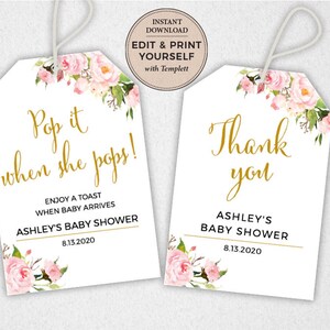 May include: Two white gift tags with pink floral designs and gold lettering. The first tag says "Pop it when she pops!" and "Enjoy a toast when baby arrives Ashley's Baby Shower 8.13.2020". The second tag says "Thank you Ashley's Baby Shower 8.13.2020".