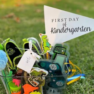 May include: A clear plastic container filled with various items for a child's first day of kindergarten. The items include a dinosaur shoe, a dinosaur eraser, a green dinosaur sticker, a blue and green game controller sticker, a blue pom-pom, a green pencil, a tag that says "WLS", and a white triangle flag with the text "FIRST DAY of kindergarten".