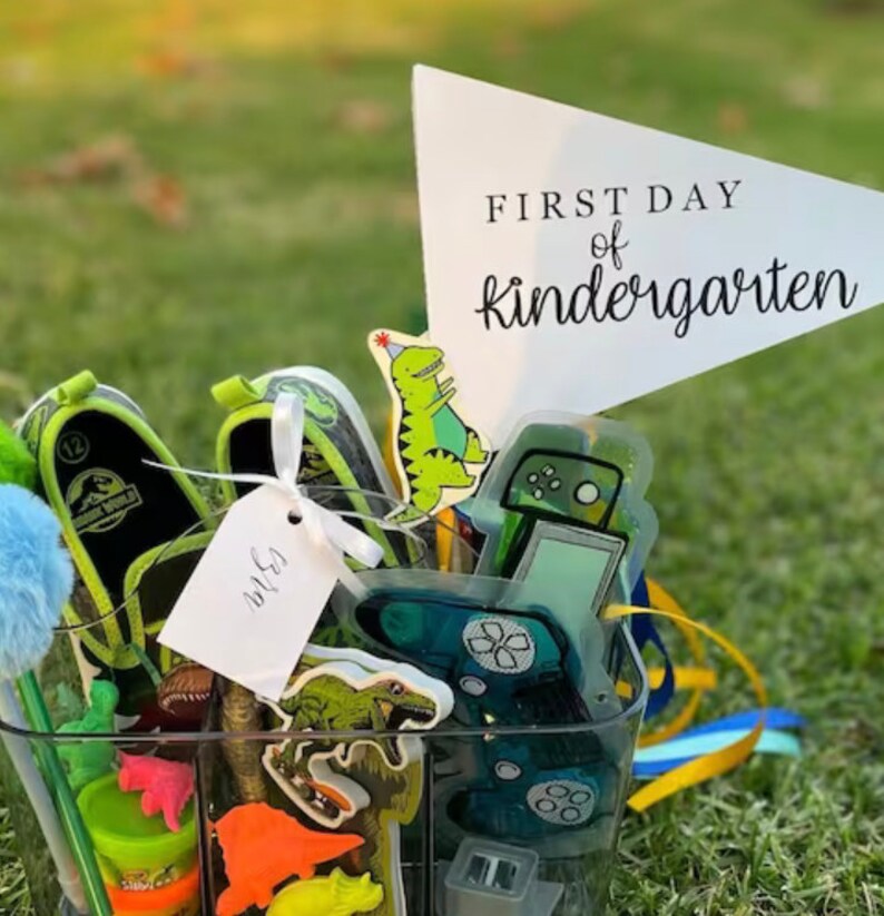 May include: A clear plastic container filled with various items for a child's first day of kindergarten. The items include a dinosaur shoe, a dinosaur eraser, a green dinosaur sticker, a blue and green game controller sticker, a blue pom-pom, a green pencil, a tag that says "WLS", and a white triangle flag with the text "FIRST DAY of kindergarten".
