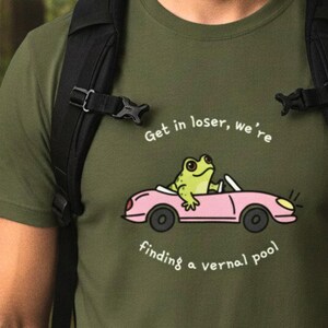 Może przedstawiać: Oliwkowozielony t-shirt z grafiką jasnoróżowego kabrioletu prowadzonego przez zieloną żabę. Tekst nad samochodem brzmi "Get in loser, we're", a tekst pod samochodem "finding a vernal pool", napisany białymi literami.