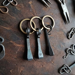 May include: Three dark gray, hand-forged metal nail pendants attached to circular brass key rings. The metal pieces have a textured, rustic finish with a visible stamped mark on the center piece. Metal pliers and additional metal links are scattered.