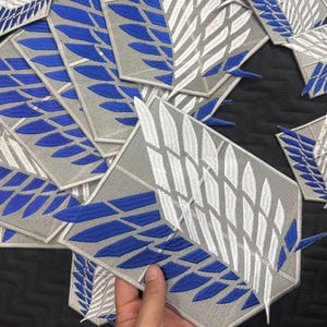 Pode incluir: Uma coleção de emblemas bordados em forma de escudo com um design de duas asas sobrepostas em fio branco e azul sobre um fundo cinza claro. Cada emblema apresenta um padrão simétrico de penas dispostas em uma forma de asa estilizada.