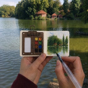 Op de afbeelding: Een klein, open schetsboek met een houten palet bevestigd door een metalen clip, met acht kleurrijke aquarelverfblokjes. De rechterpagina toont een geschilderd landschap van bomen en een gebouw aan een meer, met een waterbrush pen ernaast.