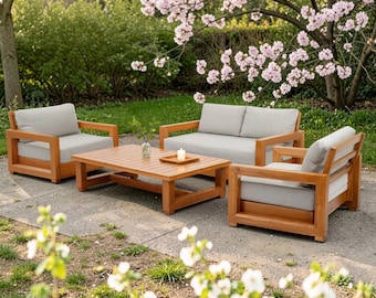 May include: A patio furniture set consisting of two armchairs, a loveseat, and a rectangular coffee table. The furniture is constructed from light brown wood with thick, light gray cushions. A small glass vase and a candle sit on the table.